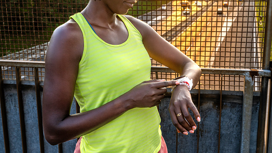 African american sportswoman checking smartwatch while jogging outdoors and standing on bridge at sunset, mid section