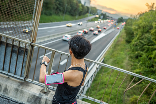 Asian sportswoman with smartphone on shoulder pocket checking smartwatch while jogging on bridge