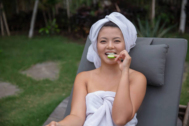 Beautiful Asian woman snacking slice of cucumber Close-up shot of Asian woman eating cucumber slice while she's sitting on a tanning bed with a wrapped towel on her head and body. woman wrapped in bath towel stock pictures, royalty-free photos & images