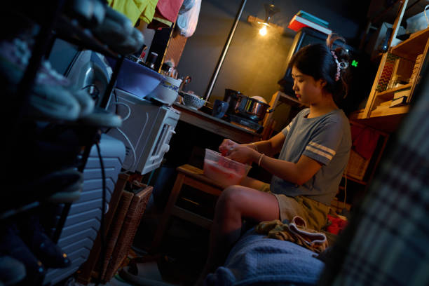 Side view of Chinese girl washing napkin or towel in plastic basin while sitting on bed against kitchen table in small apartment