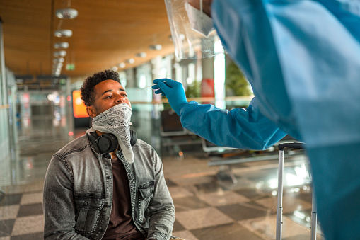 Black male traveler taking a covid-19 test at the airport terminal. Medical worker performing a nasal swab wearing protective clothes.