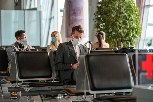 Caucasian businessman sitting in the airport lobby wearing a protective face mask while waiting to board on an airplane. Using a smartphone.