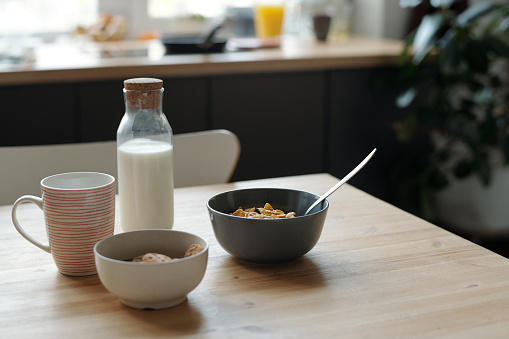 Part of wooden kitchen table with bottle of fresh milk, cup of tea or juice and two bowls with muesli or cornflakes for breakfast