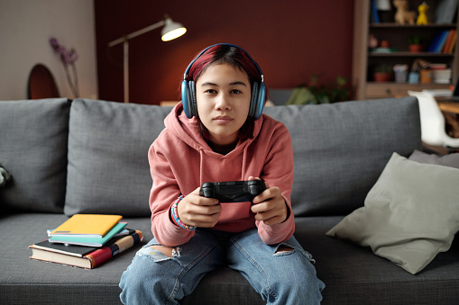 Youthful girl in headphones sitting on couch and looking at screen of computer or TV set while playing new video game at leisure