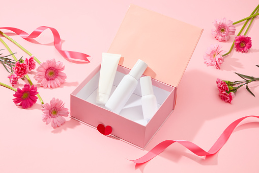Set of cosmetic bottle unlabeled placed on pink box with fresh flower (gerbera and carnation) and pink ribbon on pink background. Mockup scene for advertising with Women's day concept