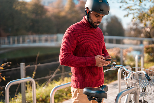 A Indian  male cyclist in a red sweater and helmet uses his smartphone while standing beside his bike in an urban park setting with a bridge in the background.
