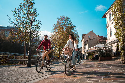 A Indian male and a Caucasian female friend enjoy a bike ride together on a sunny city street lined with trees and buildings.
