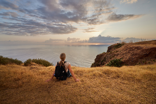 Back view of a female hiker resting on a break from her hike on cliff above the sea and looking at sunset view. Copy space.