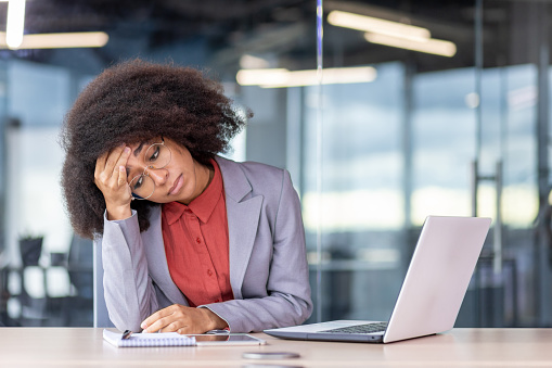 Frustrated and tired depressed business woman sitting at workplace inside office, unhappy african american woman in business suit working with laptop, failed and bankrupt.