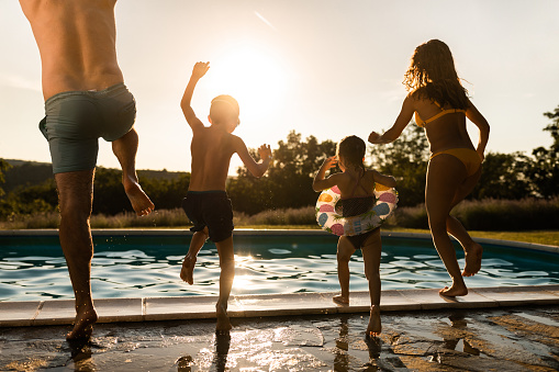 Rear view of playful parents and their kids having fun while jumping into the pool during summer sunset at the backyard. Copy space.