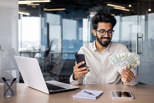 Young happy Indian man sitting in the office at the desk, holding the phone and looking at a fan of cash money.