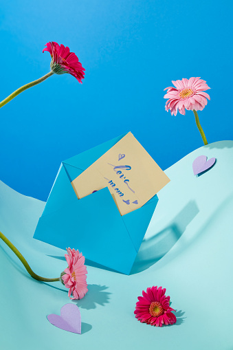 Front view of an envelope in blue color with a card inside of the text love mom. Several red and pink flowers decorated with purple paper hearts. International Womens Day is a special day