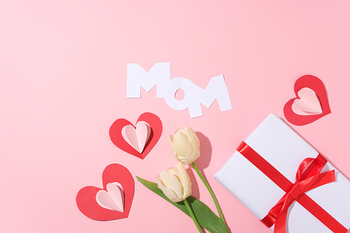 Lovely paper cut in heart shaped and Mom word arranged with tulip flowers and a gift box. Pink surface. International Women's Day is a global holiday celebrated annually on March 8