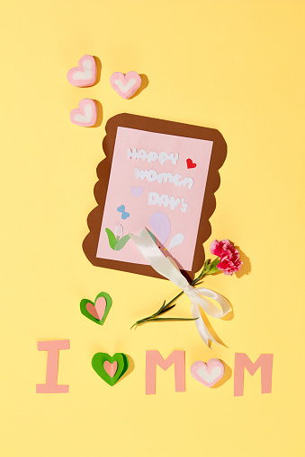 HAPPY WOMEN'S DAY text and cute sticker on a card. Heart-shaped marshmallows, a carnation and the words I LOVE MOM are decorated on a yellow background.