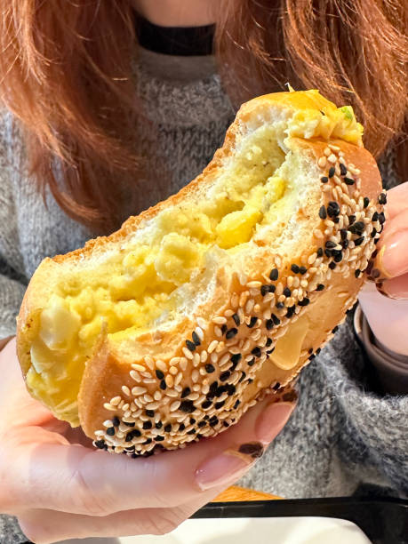 Stock photo showing close-up view of a restaurant breakfast of toasted, white bagel topped with white and black sesame seeds, filled with scrambled eggs. The sandwich can be seen, with missing bites, being held by an unrecognisable, redheaded woman with leopard skin print pattern nail art manicure. close-up image of unrecognisable, red haired woman holding toasted, white bagel with black and white sesame seeds filled with scrambled egg, missing bites, hand displaying leopard skin print pattern nail art manicure , focus on foreground