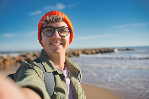 Young man with cap and glasses taking a selfie on a winter holiday - Happy hiker with backpack smiling at the camera outdoors - Tourist walking on the beach - Travel and technology.