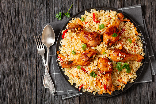 fried chicken drumsticks over brown rice pilaf with vegetables and raisins on black platter on dark wooden table with spoon and fork, horizontal view from above, flat lay, free space