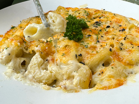 Stock photo showing close-up, elevated view of a white bowl containing macaroni and cheese / Mac and cheese baked pasta in white cheese sauce with garnish of parsley.