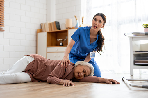 Asian caregiver help senior female from falling on the ground at home. Elderly older patient having an accident while doing physical therapy then rescued by attractive therapist nurse in living room.