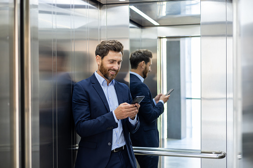 Confident businessman in a suit smiling at his phone in an elevator, reflecting success and connectivity.