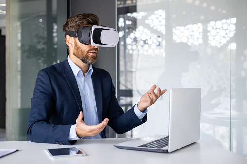 Businessman in suit using VR headset at desk in modern office, gesturing with hands, exploring virtual reality.