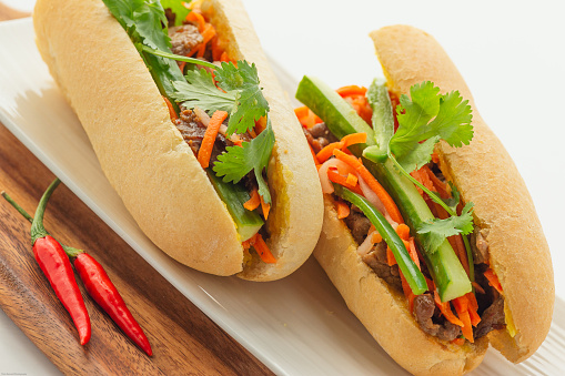 Cut up Vietnamese satay beef baguette with variety of veggie on wooden cutting board on white background