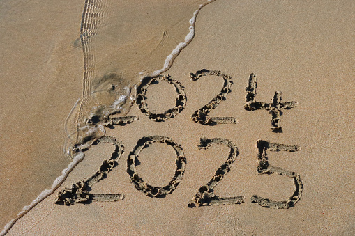 Stock photo showing close-up view of the year dates 2024 and 2025 written in the sand on a sunny beach by the sea's water's edge at low tide with threat of being wiped away. New Year's in southern hemisphere concept.