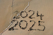 Close-up image of writing drawn on sunny beach with year dates 2024 and 2025 written in sand with stick, water's edge of sea at low tide, handwriting numbers in soft golden sand, elevated view, New Year's in southern hemisphere concept