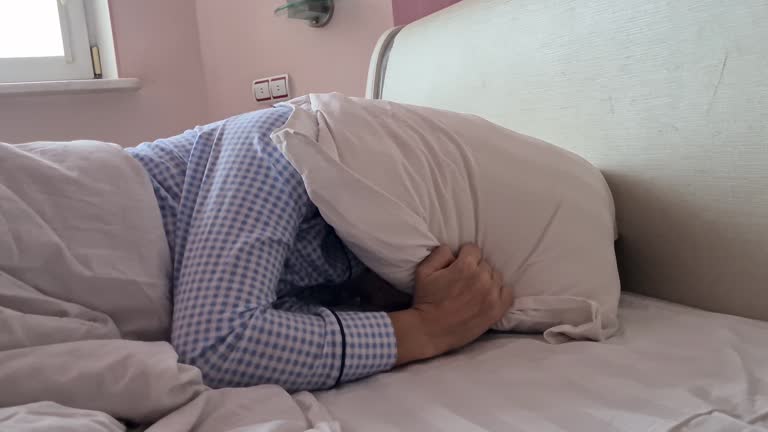 Young woman lying in bed at home and covering her ears with pillow and trying to sleep