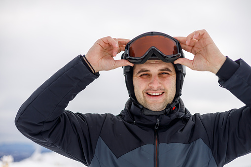 The photo shows a young skier, standing confidently on a snowy hill, looking directly at the camera. His attire, complete with ski poles and goggles, highlights his readiness to tackle the slopes and enjoy the cold, picturesque landscape