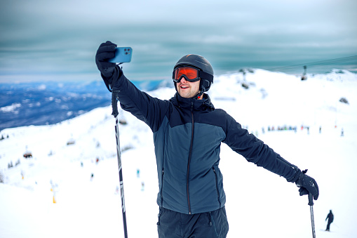 A young man pauses on the ski slopes to capture a selfie, his smartphone in hand against the stunning backdrop of snow-covered mountains, merging the thrill of skiing with the instant shareability of modern technology