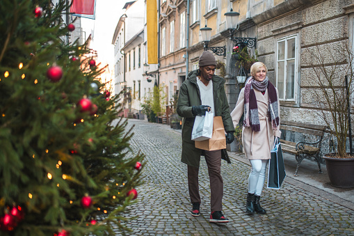 A mixed race young adult male and a Caucasian  female stroll together along a cobblestone street by a festive Christmas tree, enjoying a winter day shopping with bags in hand.