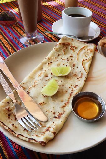 Stock photo showing close-up, elevated view of a light, healthy breakfast consisting of a folded crepe with lemon wedges and a metal ramekin of honey.