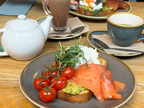 Stock photo showing close-up view of a plate containing a slice of white toasted bread topped with mashed avocado, grilled cherry tomatoes on the vine, poached egg, smoked salmon, garnished with pea shoots,  besides a white teapot, teacup and sauce and a drinking glass of hot chocolate, in a restaurant setting.