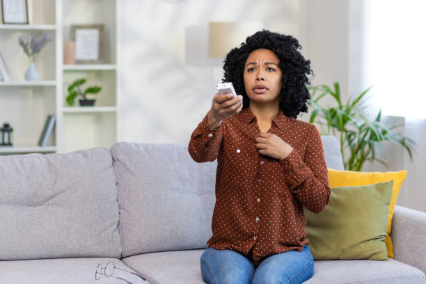 besorgte junge afroamerikanerin, die zu hause auf dem sofa sitzt und die klimaanlage per fernbedienung einschaltet. er fühlt sich schlecht - air conditioner cold home interior climate stock-fotos und bilder