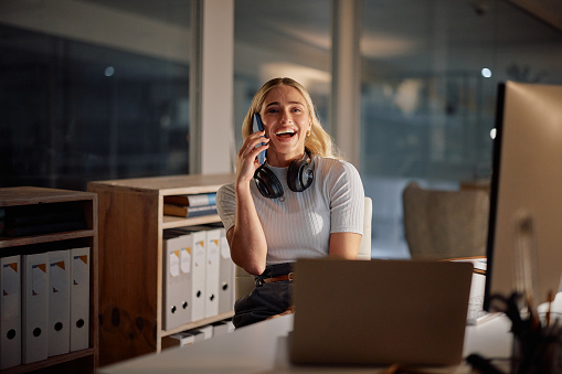 Young businesswoman, phone call and office at night by computer, laughing and gossip chat for relax. Producer, headphones and smartphone at desk in discussion, funny joke and wellness in workplace