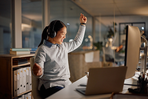 Night in office, woman with headphones and dancing at desk with smile, computer and tech startup. Late shift, music and happy celebration, fun and streaming for Asian girl in workplace with laptop.