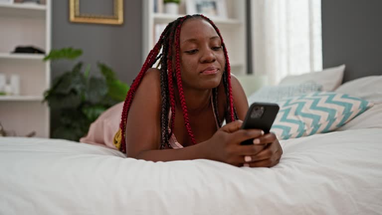 Surprised african american woman in pajamas using phone in bedroom bed