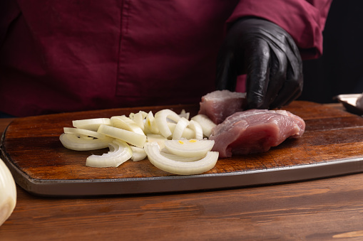 A cook wearing black gloves is slicing onions into half rings and cutting raw pork meat. On the chopping board, there are raw meat and onions