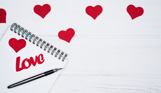 Flatlay composition with notebook, pen and red hearts on a white wooden background. Romantic congratulations to Valentine's Day. Top view. Place for text.