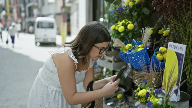 Beautiful hispanic woman in glasses snapping pictures of vibrant flowers on tokyo's streets