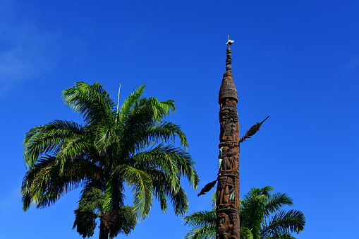 Nouméa, South Province, New Caledonia: Kanak totem and 'fleche faitiére' (rooftop arrow) on Mwa Ka / Mwaka Square, downtown Nouméa - an indigenous Melanesian symbol that came to represent the Kanak people and the independence movement (FLNKS).  A respected emblem of the Kanak chiefdom, the 'fleche faitiére', is a traditional element of Kanak architecture, which dominates and adorns the cone-shaped thatched roof of the large huts and ceremonial totems of a clan. It is made with houp (Montrouziera cauliflora), a rot-proof wood from high-altitude forests and has a a calling conch at the top.