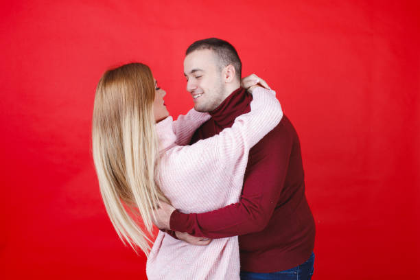 Young heterosexual couple in turtleneck sweaters hugging and smiling at each other on Valentine's Day, red background, studio shot