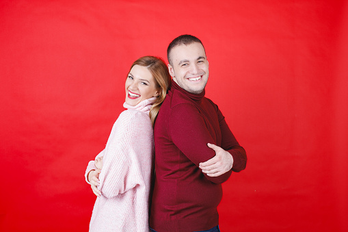 Young couple in turtleneck sweaters standing back to back and smiling at camera with joy on Valentine's Day, red background, studio shot