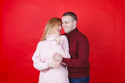 Young man embracing his girlfriend affectionately from behind, she is smiling joyfully on Valentine's Day, red background, studio shot