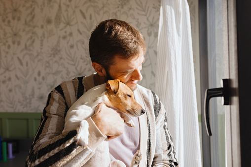 Man hugging his cute little dog while looking through the window