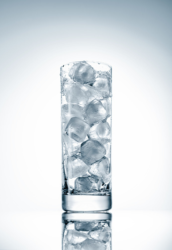 A tall transparent glass cup filled with natural ice cubes on a light background, backlit, studio shot.