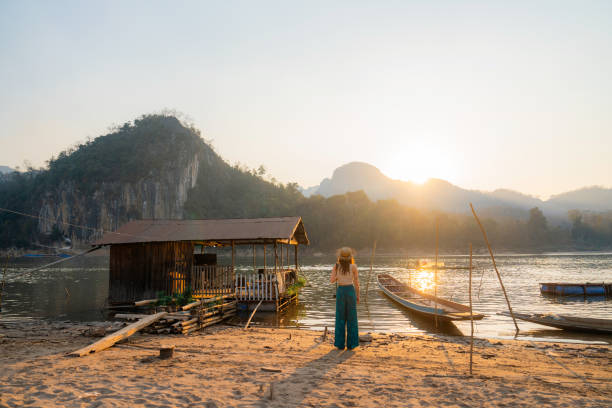 Serene woman in straw hat photographing at sunset near  Mekong river during experiential travel to Laos
