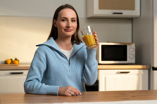 Young brunette dressed in a blue sweater drinks orange juice from a glass while sitting at the kitchen table.