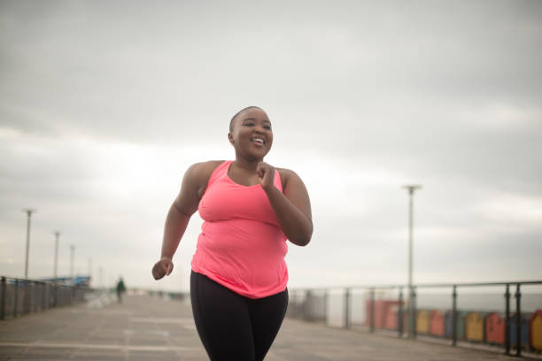 Fitness, smile and plus size black woman running on promenade at beach in morning for health, exercise or cardio. Marathon, sports and happy young runner or athlete outdoor for training or workout stock photo